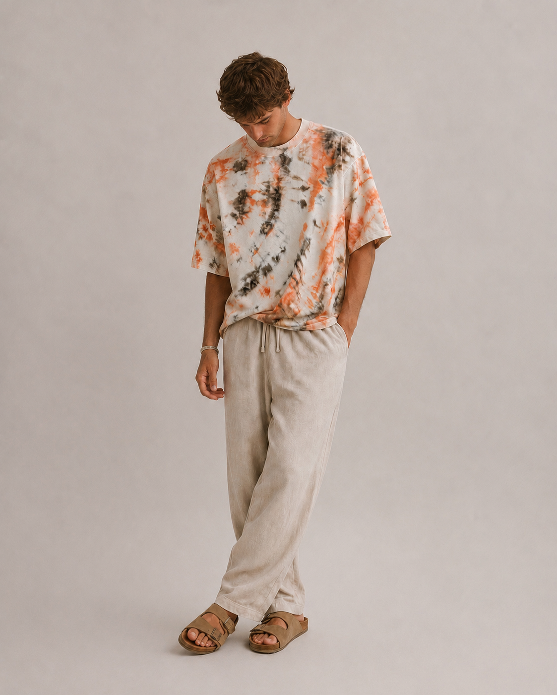 A man wearing a short-sleeve tie-dye t-shirt in orange, brown, paired with light beige Hemp drawstring pants and brown sandals, standing against a neutral light gray studio background. Hemp Horizon