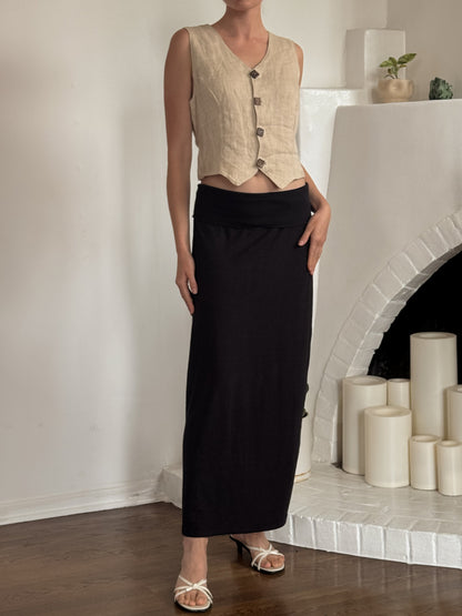 A model stands in a bright room wearing a sleeveless beige linen vest with dark buttons and a long black skirt. The background features a white brick fireplace with white candles.