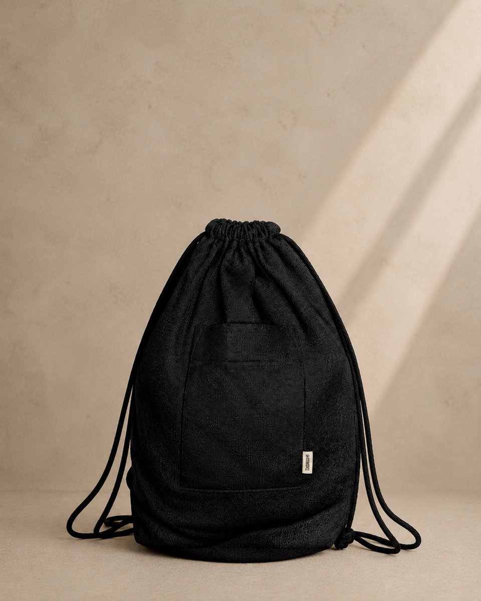 A black drawstring backpack made of textured canvas, featuring a front pocket with a small white brand tag, set against a warm beige textured wall with soft diagonal shadows.