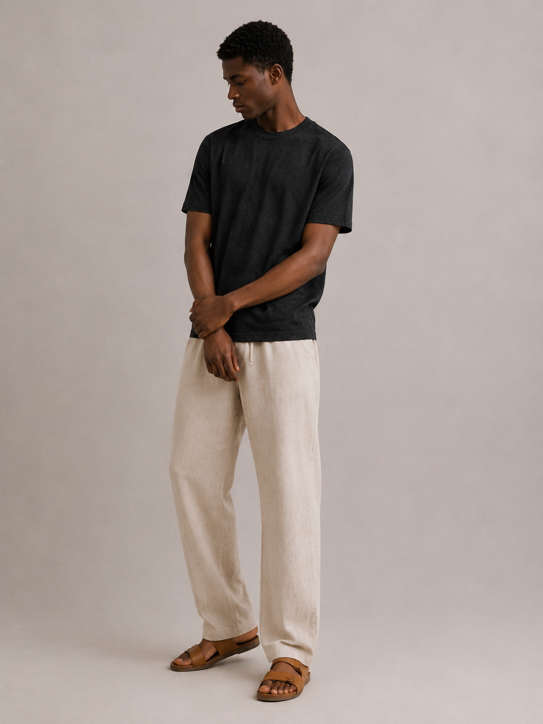 A man modeling a black short-sleeve Hemp and organic cotton t-shirt and off-white linen trousers, paired with brown leather slide sandals, against a neutral light gray studio background. Hemp Horizon