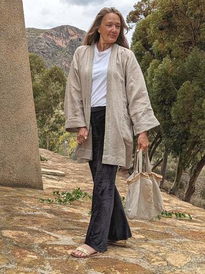 A person wearing a beige hemp kimono dress jacket with a belt, walking down stone stairs. They are also carrying a beige bag and wearing sneakers.