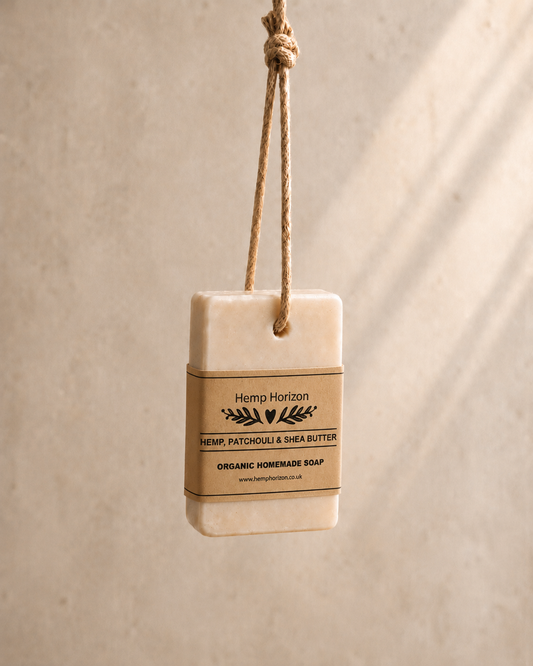 A rectangular bar of Hemp Horizon Hemp, Patchouli & Shea Butter organic homemade soap hangs from a natural jute rope tag against a textured beige wall with soft diagonal shadows. Hemp Horizon