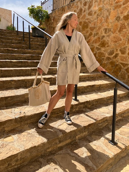 A person wearing a beige hemp kimono dress jacket with a belt, walking down stone stairs. They are also carrying a beige bag and wearing sneakers.