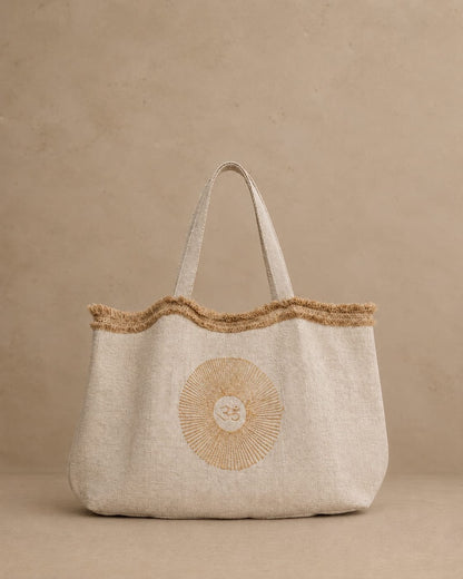 A natural hemp tote bag with a frayed jute trim along the top edge and double shoulder straps and internal pocket, featuring a circular OM motif block printed on the front, set against a textured beige background.