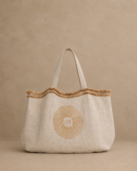 A natural hemp tote bag with a frayed jute trim along the top edge and double shoulder straps and internal pocket, featuring a circular OM motif block printed on the front, set against a textured beige background.