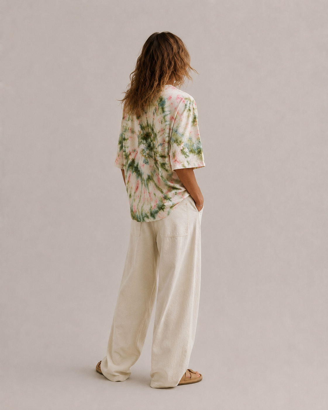 A woman is shown from behind wearing a short-sleeve ice-dye t-shirt with green, pink, and white swirls, paired with wide-leg hemp cream pants and tan sandals, standing against a neutral beige studio background. Hemp Horizon