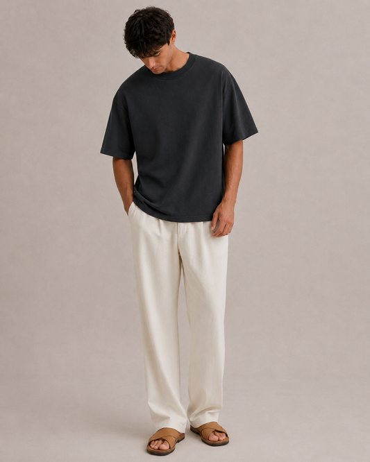 A man wearing a black oversized Hemp and Bamboo t-shirt, with off-white wide-leg trousers, and brown leather slide sandals, standing against a neutral beige studio background. Hemp Horizon