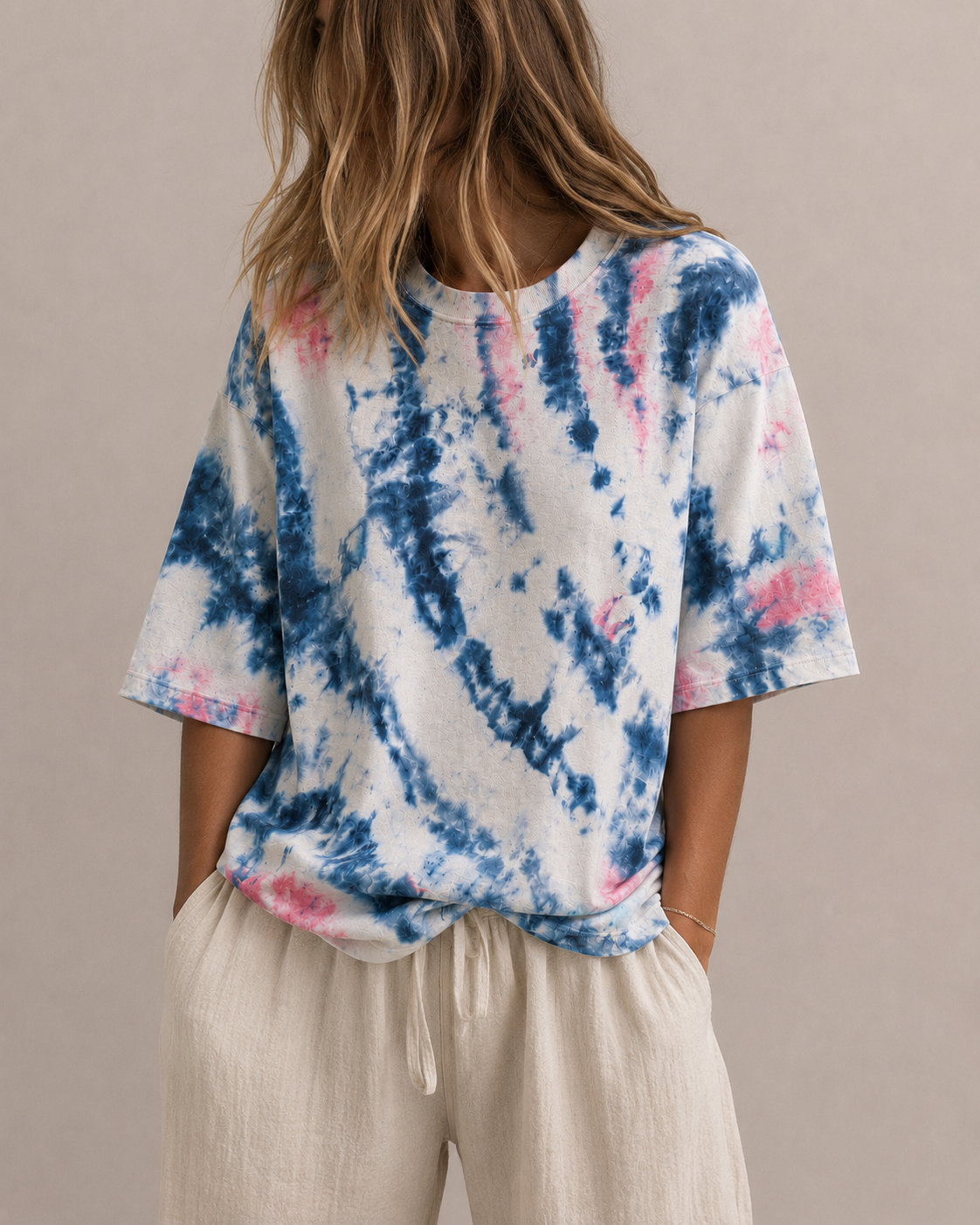 A woman wearing a short-sleeve tie-dye t-shirt with blue, pink, and natural patterns, paired with light beige hemp pants, shown from a rear three-quarter view against a solid beige background. Hemp Horizon