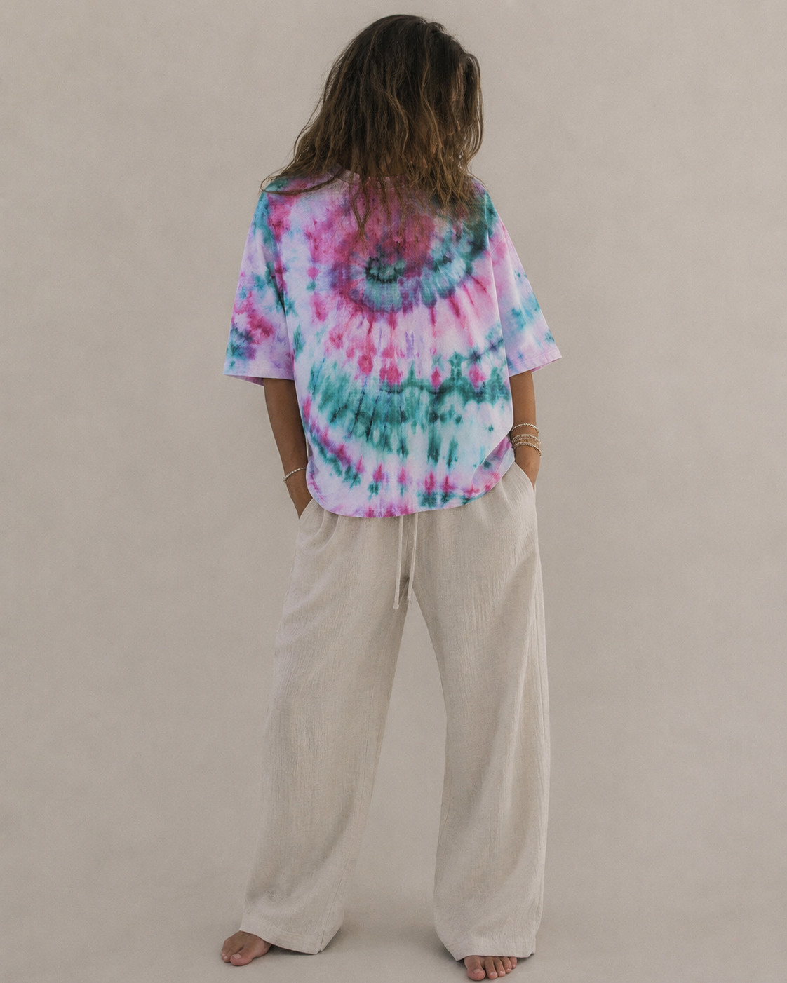A woman shown wearing a colorful turquoise and pink tie-dye t-shirt and cream-colored wide-leg pants against a plain hemp beige background. Hemp Horizon