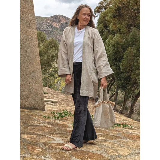 A person wearing a beige hemp kimono dress jacket with a belt, walking down stone stairs. They are also carrying a beige bag and wearing sneakers.