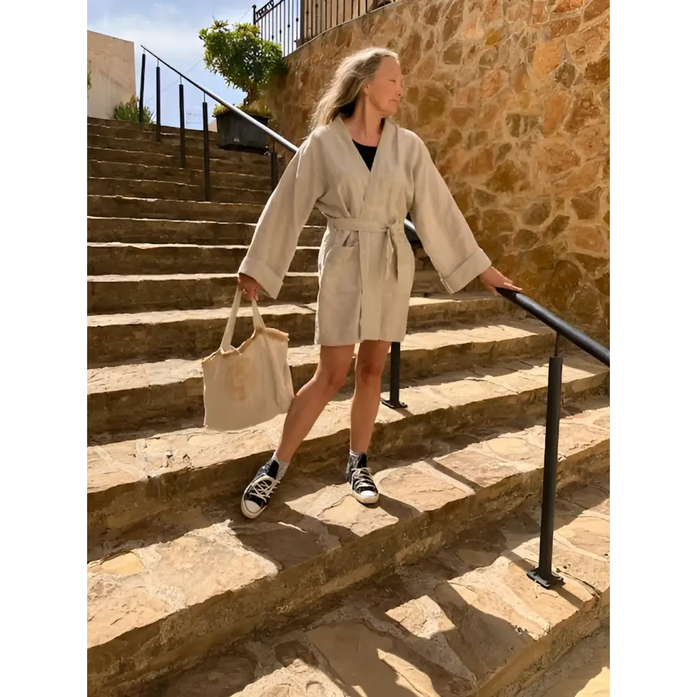 A person wearing a beige hemp kimono dress jacket with a belt, walking down stone stairs. They are also carrying a beige bag and wearing sneakers.