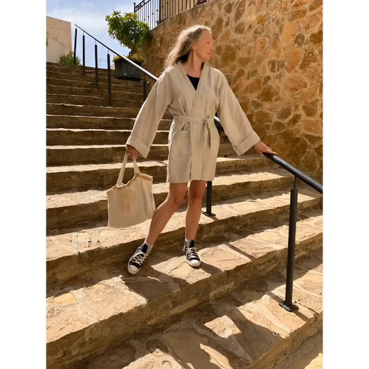 A person wearing a beige hemp kimono dress jacket with a belt, walking down stone stairs. They are also carrying a beige bag and wearing sneakers.